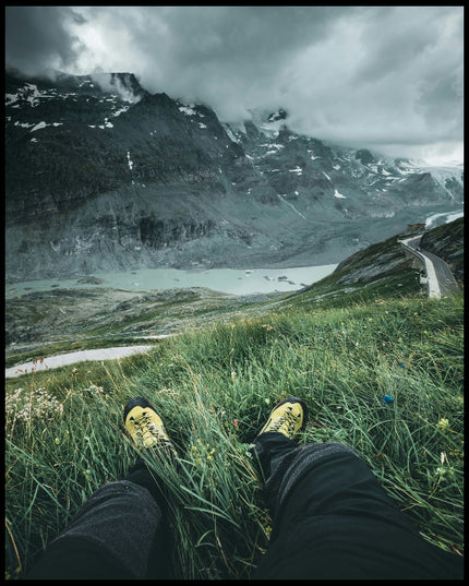 Leinwandbild aus der Egoperspektive von einer Person die im Gras sitzt und auf eine Berglandschaft blickt.