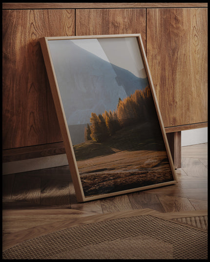 Ein gerahmtes Poster von einer Herbstlandschaft in den Dolomiten mit Bäumen im Sonnenlicht, an einer Holzkommode angelehnt und stehend auf dem Holzboden