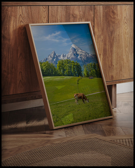 Ein gerahmtes Poster von einer Kuh auf einer grünen Wiese vor einer Alpenlandschaft, an einer Holzkommode angelehnt und stehend auf dem Holzboden