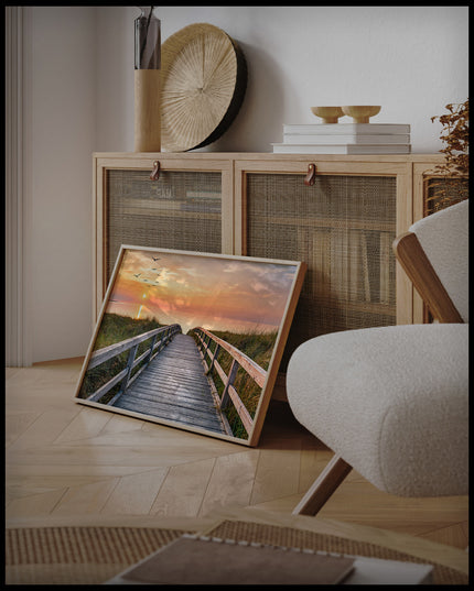 Ein gerahmtes Poster von einem Holzsteg im Sonnenuntergang, der über Dünen zum Strand führt, an einer Holzkommode angelehnt und stehend auf dem Holzboden