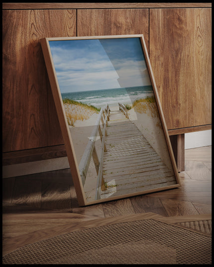 Ein gerahmtes Poster von einem Steg aus Holz der durch sandige Dünen zum Strand führt, an einer Holzkommode angelehnt und stehend auf dem Holzboden