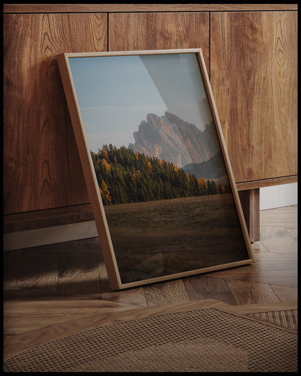 Ein gerahmtes Poster von einem dichten Waldstück in einer kargen Berglandschaft, an einer Holzkommode angelehnt und stehend auf dem Holzboden