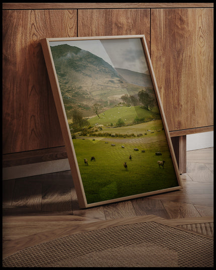 Ein gerahmtes Poster von einer grünen Weide mit Schafen vor einem Berg, an einer Holzkommode angelehnt und stehend auf dem Holzboden