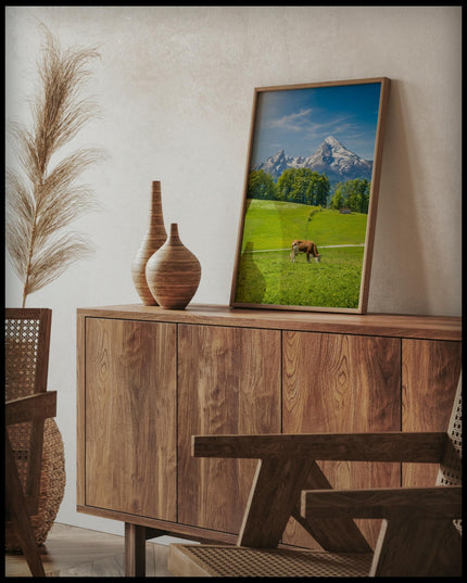 Ein gerahmtes Poster von einer Kuh auf einer grünen Wiese vor einer Alpenlandschaft, auf einer Holzkommode an die Wand angelehnt