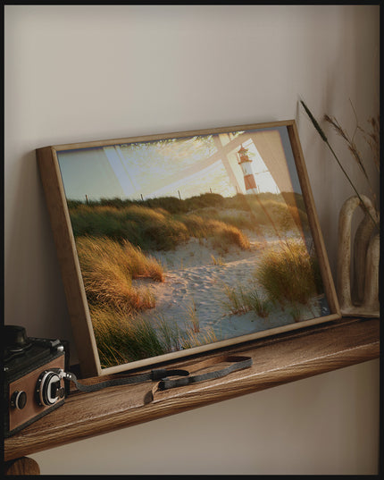 Ein gerahmtes Poster von einem Leuchtturm auf Sylt, der hinter grasbedeckten Dünen hervorschaut, auf einem Holzregal an die Wand angelehnt