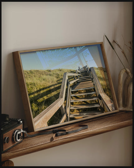 Ein gerahmtes Poster von einer Holztreppe die über Dünen am Strand führt, auf einem Holzregal an die Wand angelehnt