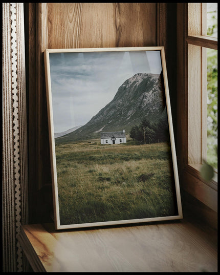 Ein gerahmtes Poster von einer weißen Holzhütte auf einer Wiese vor einem Berg, vor einem Fenster an die Wand angelehnt