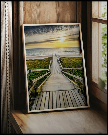 Ein gerahmtes Poster von einem Holzsteg mit einer Treppe, der durch Dünen zum Strand führt, vor einem Fenster an die Wand angelehnt