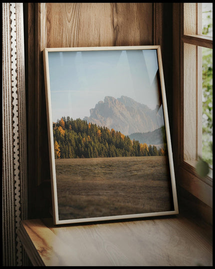 Ein gerahmtes Poster von einem dichten Waldstück in einer kargen Berglandschaft, vor einem Fenster an die Wand angelehnt