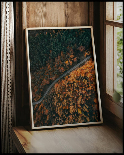 Ein gerahmtes Poster aus der Vogelperspektive von einer Straße die durch einen herbstlichen Wald führt, vor einem Fenster an die Wand angelehnt